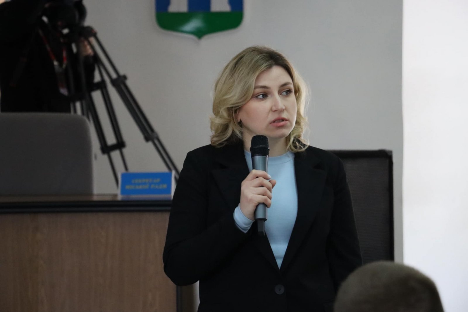 Жінка у темному костюмі з мікрофоном стоїть перед аудиторією