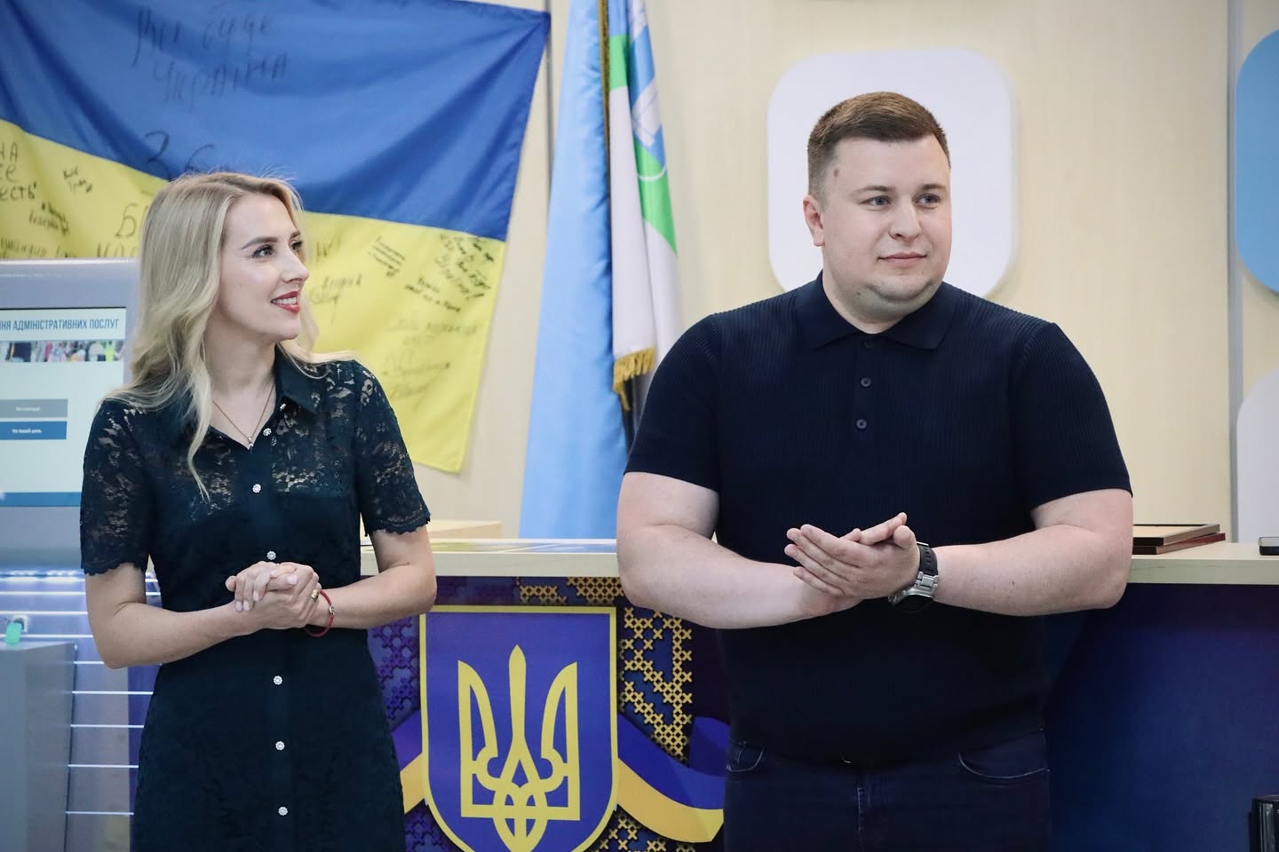 Чоловік і жінка стоять біля стійки з прапорами, аплодують і усміхаються.