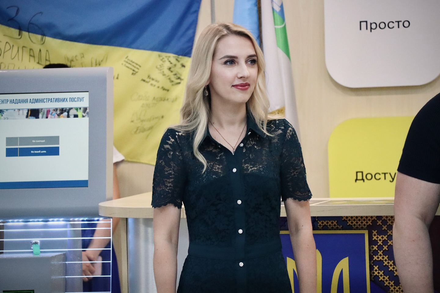 Жінка у темній сукні стоїть у залі, позаду прапор України.