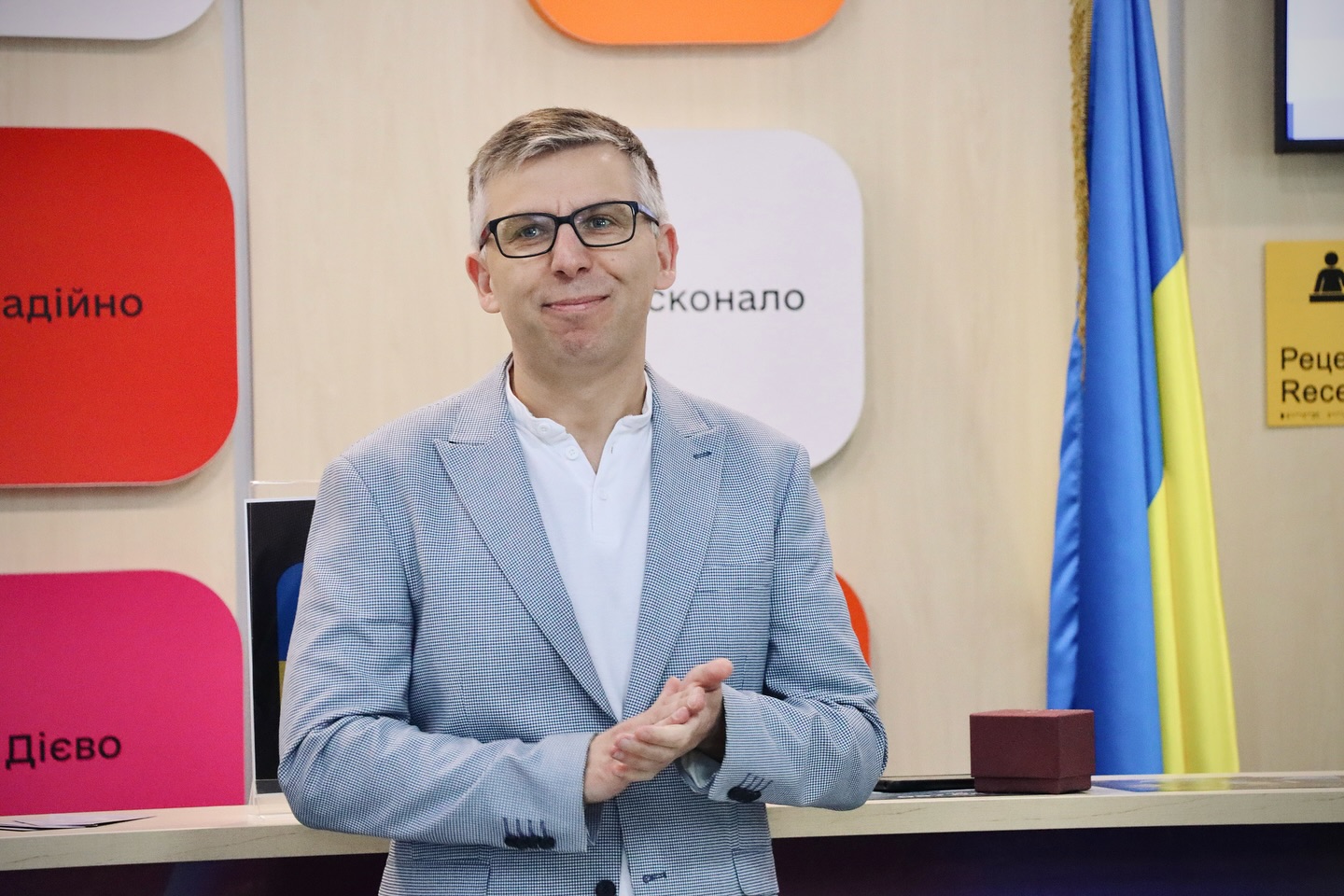 Чоловік у сірому піджаку стоїть біля стійки, аплодує, усміхаючись.