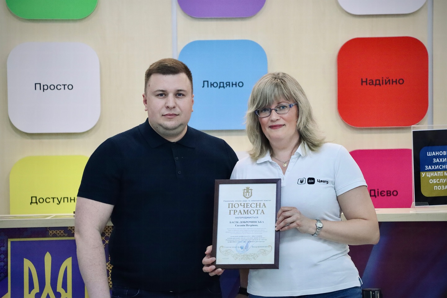 Чоловік і жінка тримають почесну грамоту у приміщенні ЦНАП, дивляться в камеру.