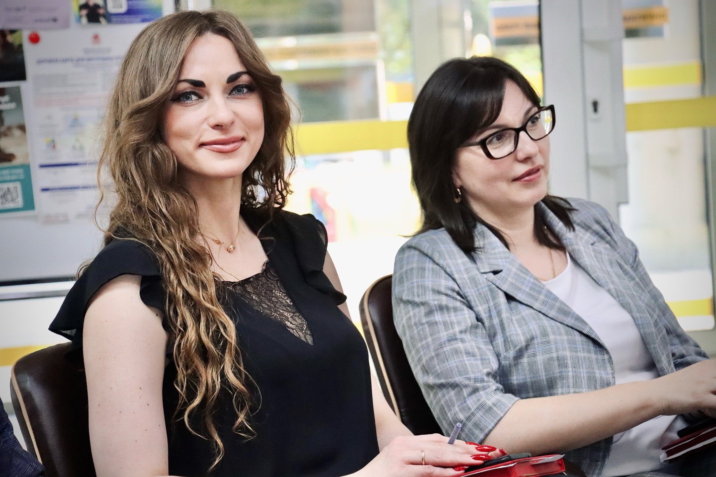 Дві жінки сидять у приміщенні та посміхаються.