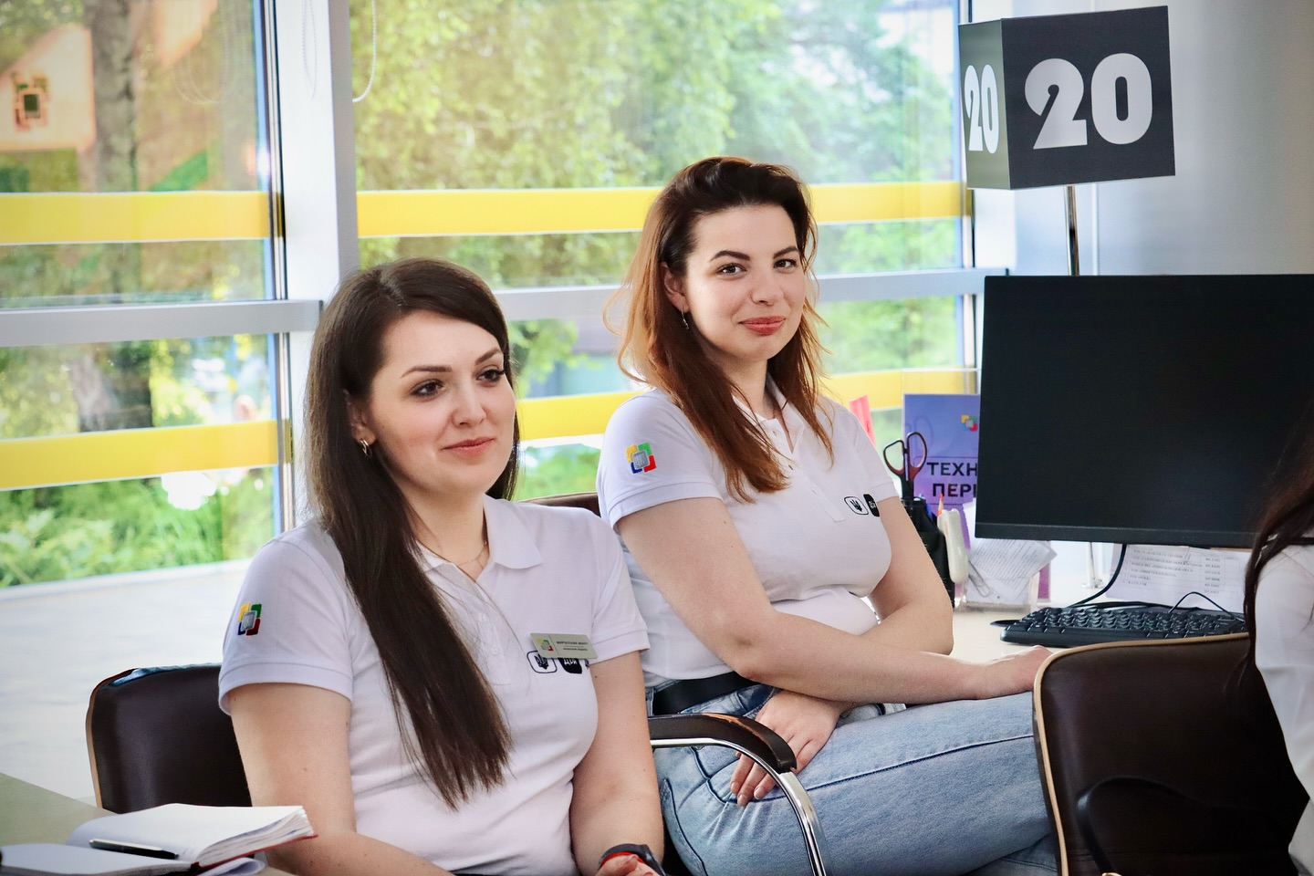 Дві жінки у білому одязі сидять у приміщенні та посміхаються.