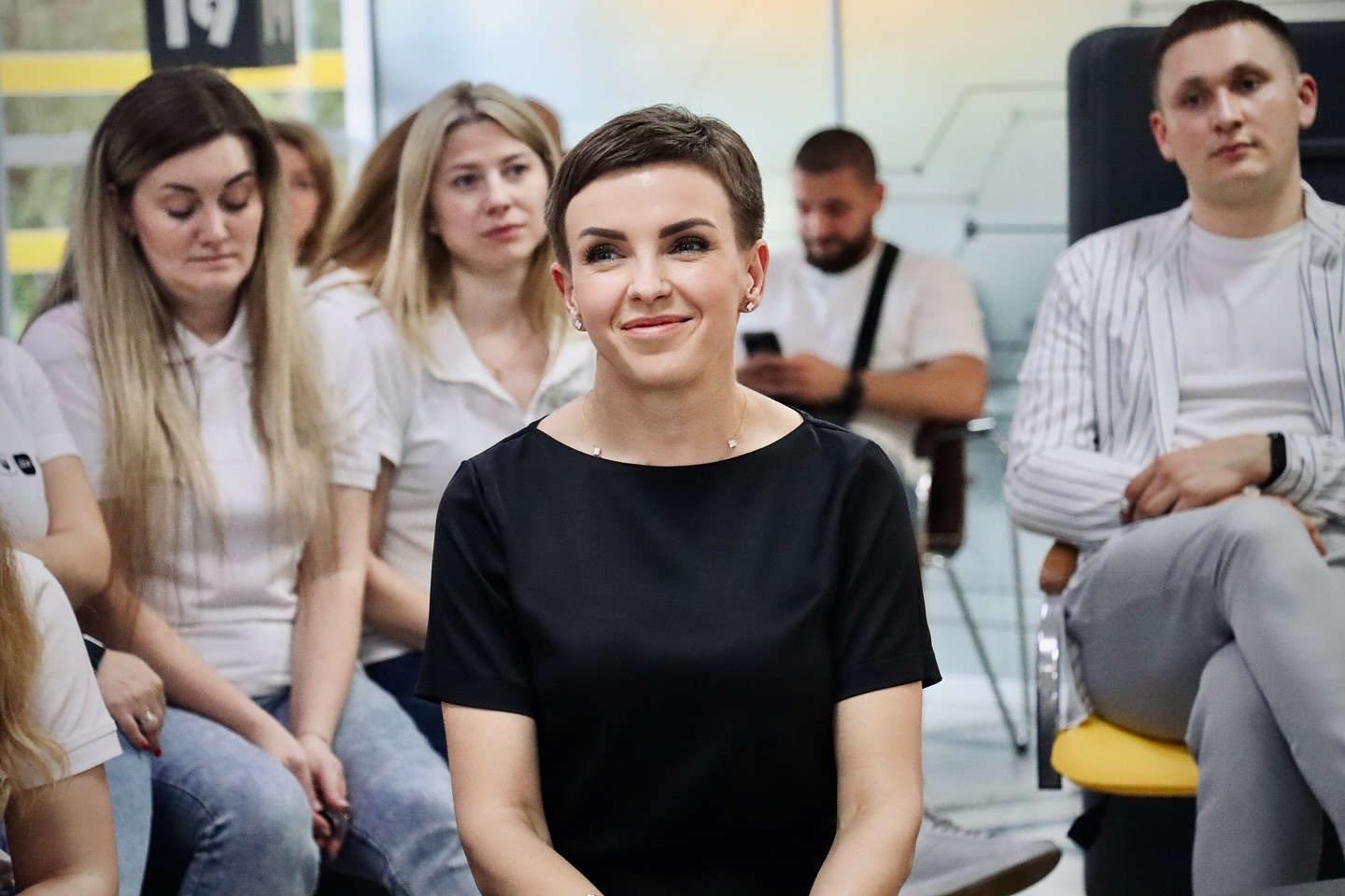 Жінка в чорному одязі сидить на заході серед інших учасників та усміхається.