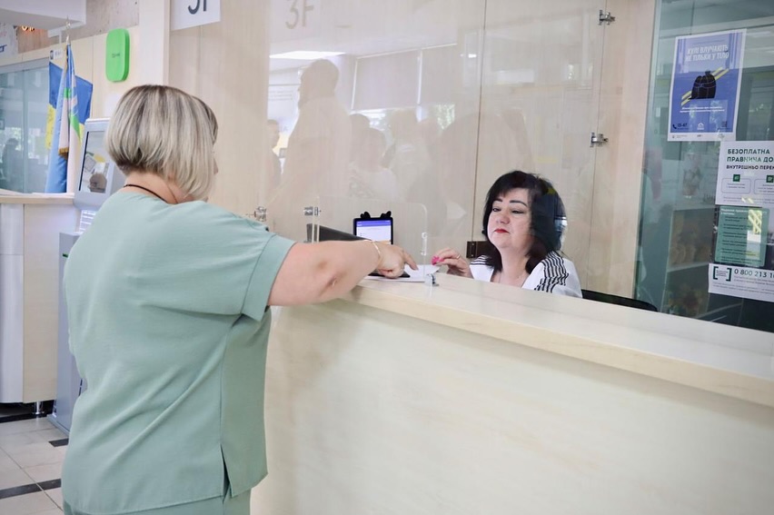 Жінка в зеленому костюмі звертається до працівниці за стійкою в установі.
