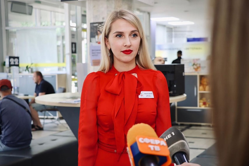 Жінка зі світлим волоссям, у червоній сукні, дає інтерв'ю у приміщенні.