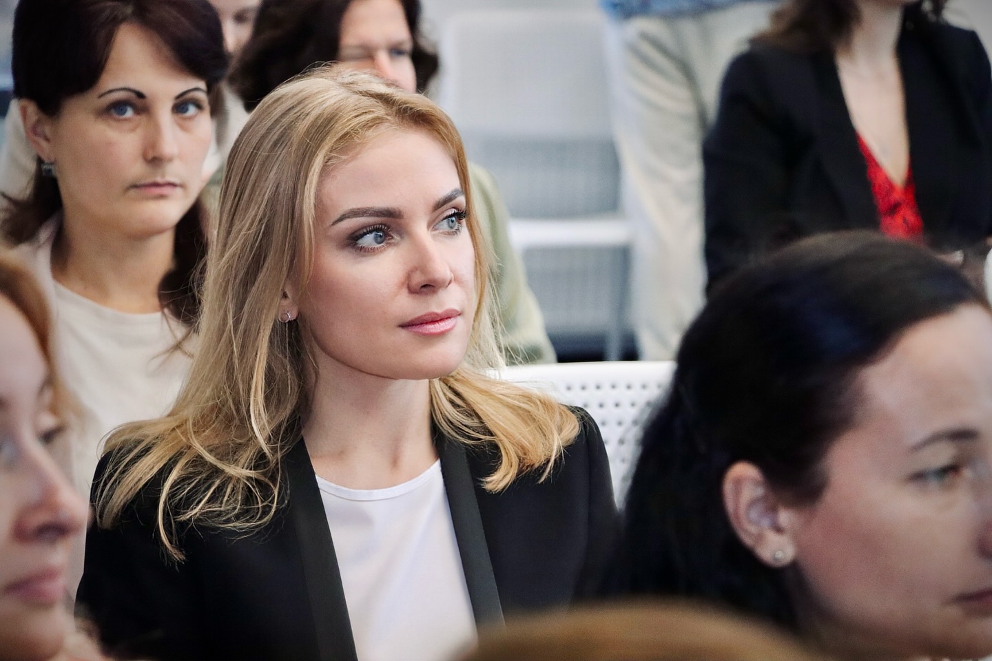Жінка із світлим волоссям сидить в приміщенні серед інших людей.