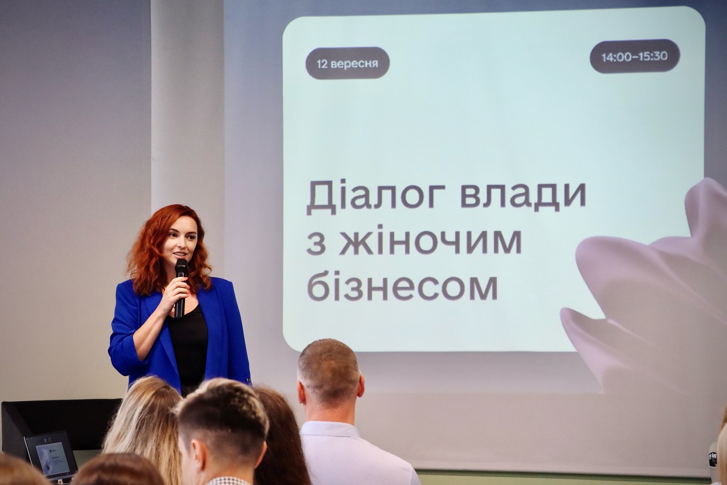 Жінка з рудим волоссям розповідає щось в мікрофон, на задньому фоні - презентація.