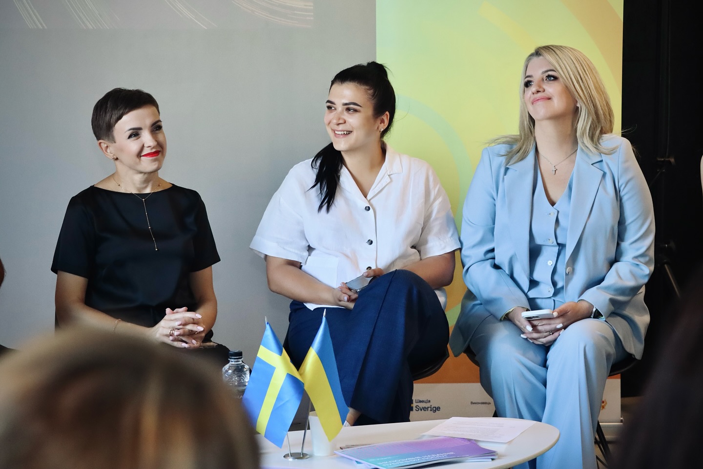 Три жінки сидять у приміщенні та посміхаються.