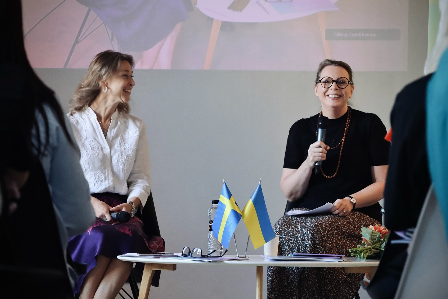 Дві жінки сидять на сцені під час заходу, одна з мікрофоном. На столику перед ними прапорці.