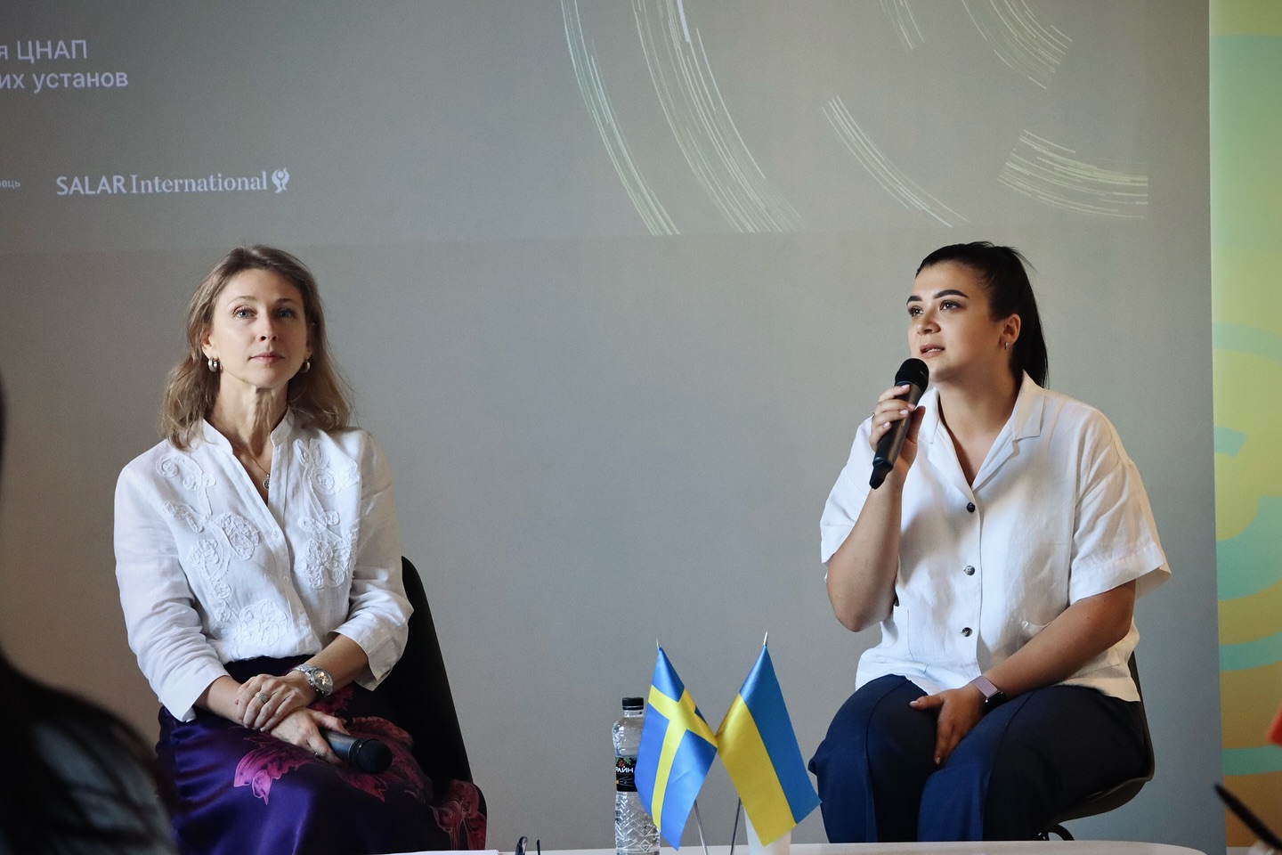 Дві жінки сидять на сцені, одна з мікрофоном. На столику перед ними — прапорці України та Швеції.