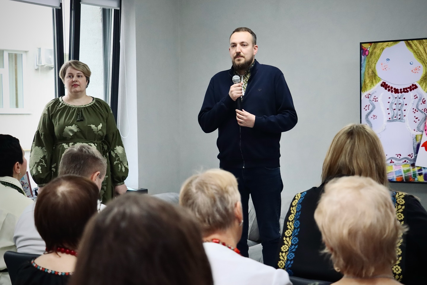 Чоловік з мікрофоном стоїть перед групою людей у приміщенні.