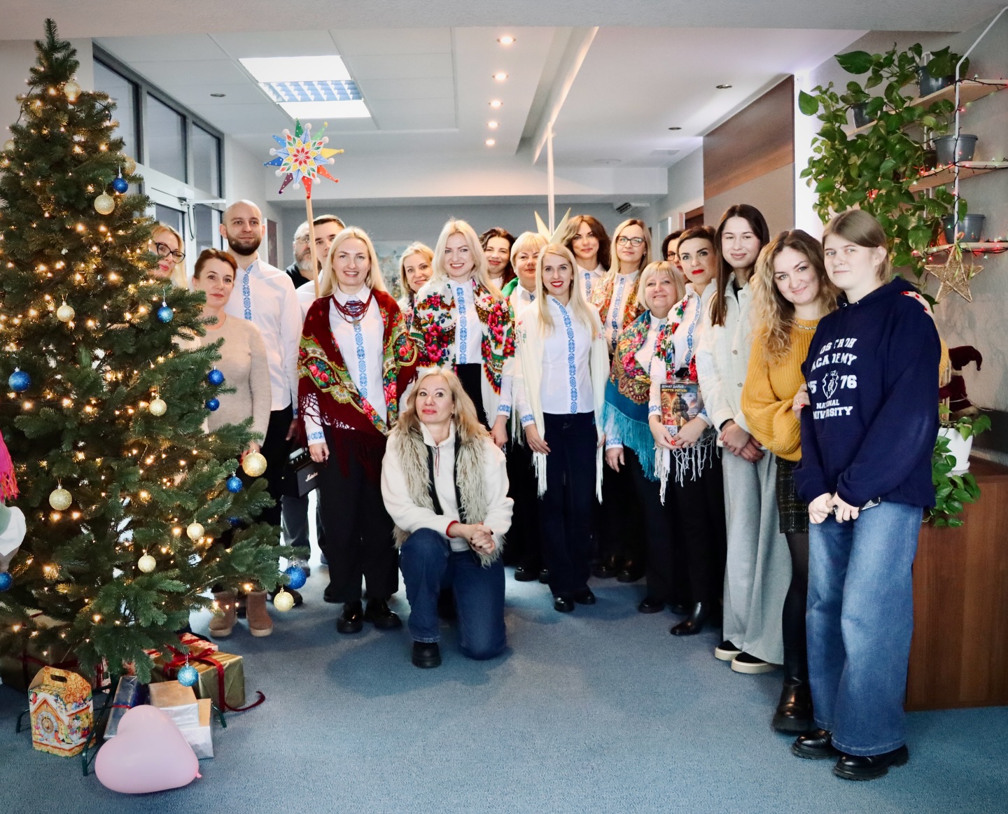 Група людей у вишитих сорочках позує для фото біля ялинки.