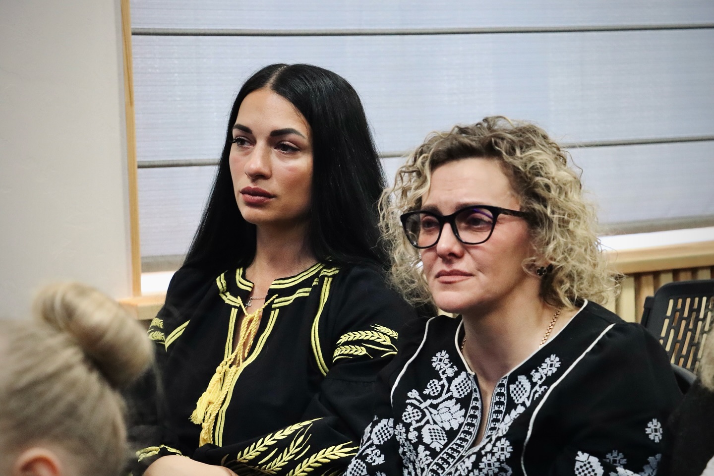 Дві жінки у чорних вишиванках сидять у приміщенні.