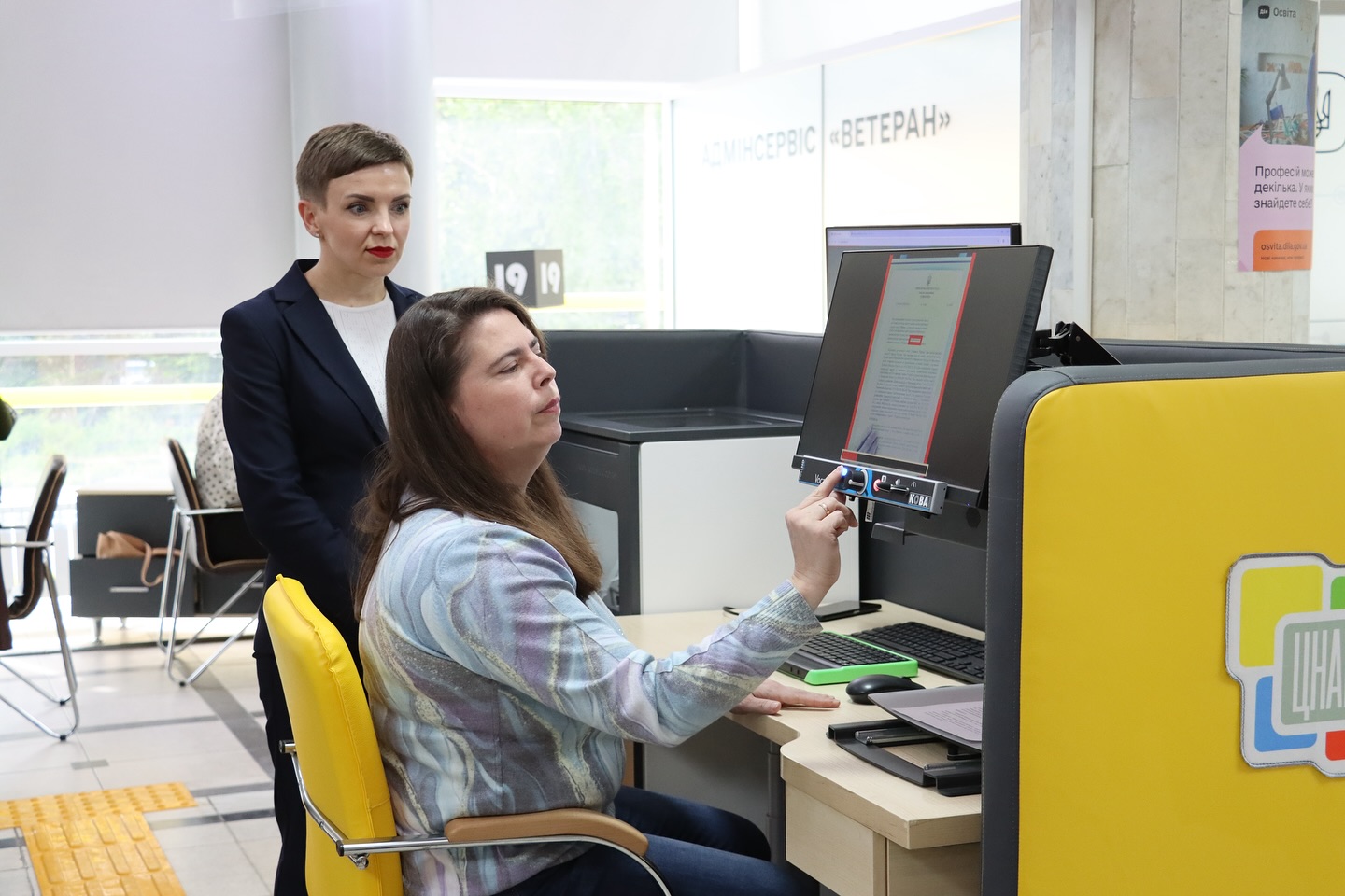 Жінка користується пристроєм для зчитування документів у приміщенні, поруч стоїть інша жінка.