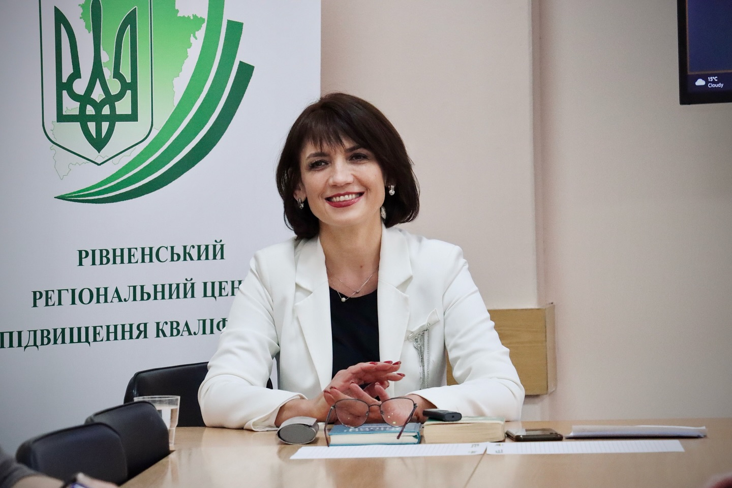 Жінка з коротким темним волоссям сидить за столом у світлому піджаку.