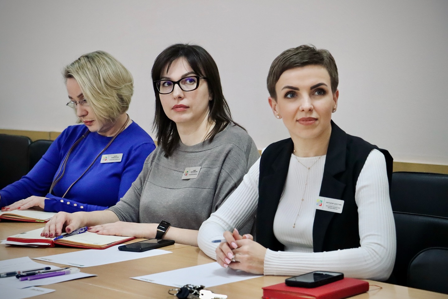 Три жінки з бейджами сидять за столом, дивлячись убік лектора.