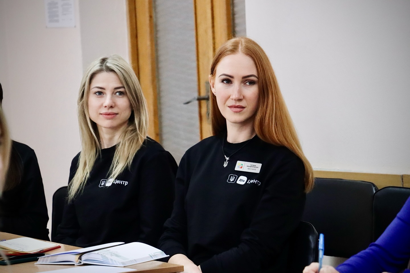 Дві жінки сидять за столом. Вони одягненні у чорні брендовані кофти. На столі лежать записники.