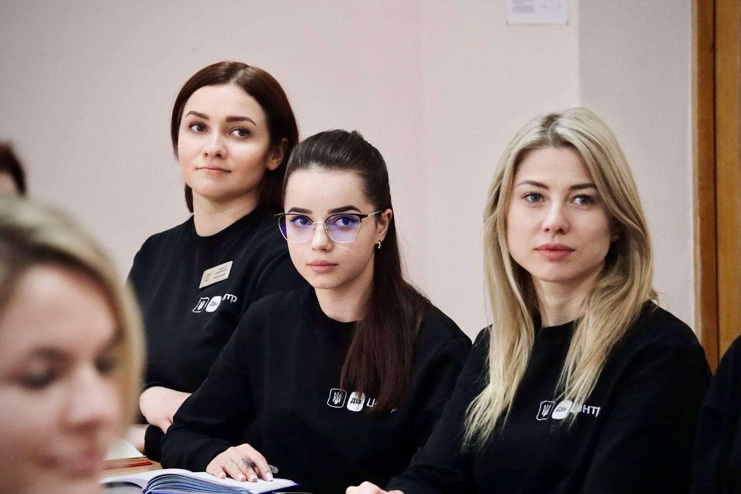 Дві жінки та дівчина в окулярах сидить за столом у чорних кофтах, уважно слухаючи лектора.
