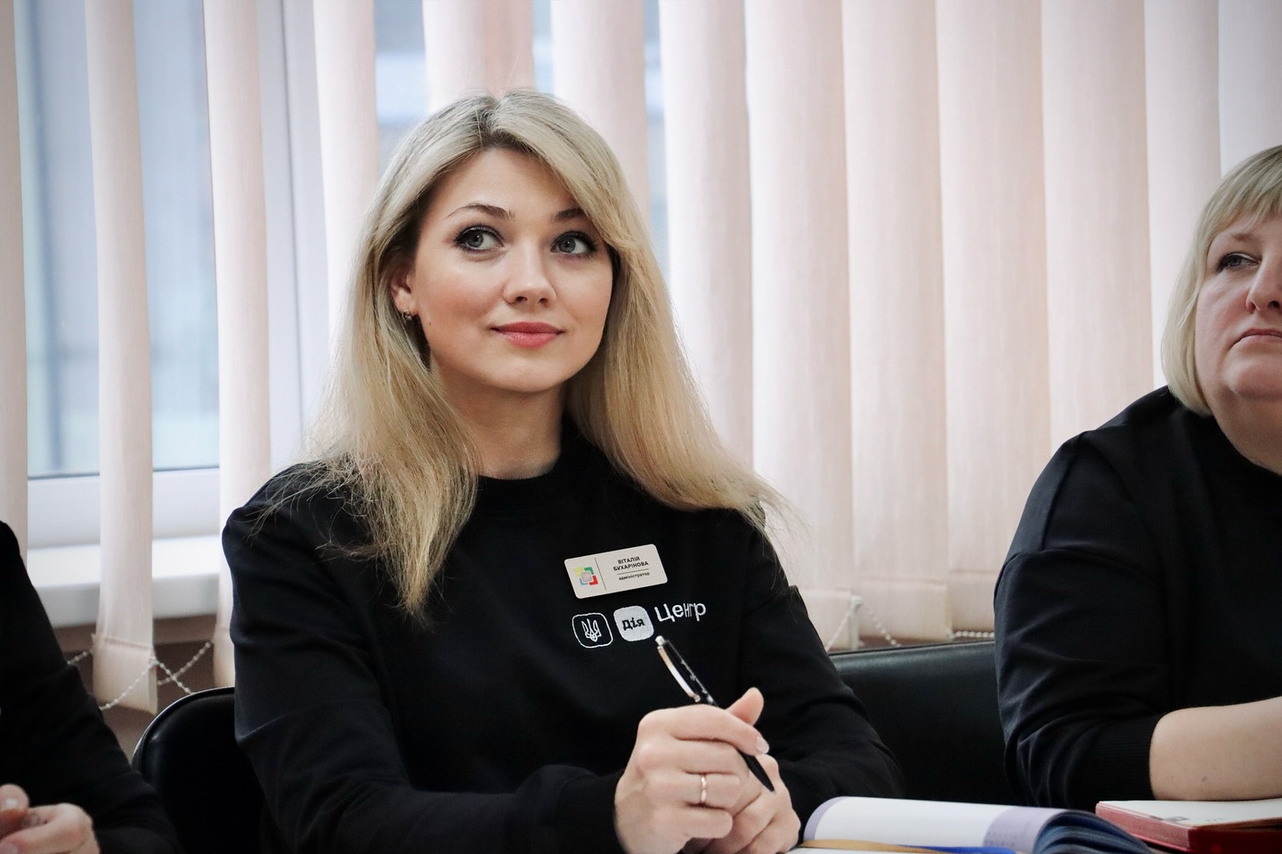 Жінка з білим волосся у чорній кофті з бейджем сидить за столом. У руках вона тримає кулькову ручку.