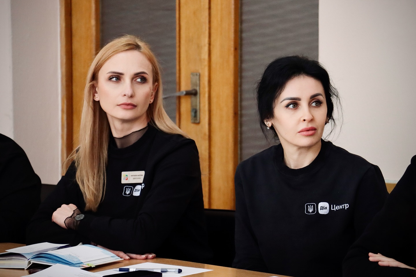 Дві жінки у чорних кофтах з бейджами сидять за столом, уважно слухаючи доповідача.
