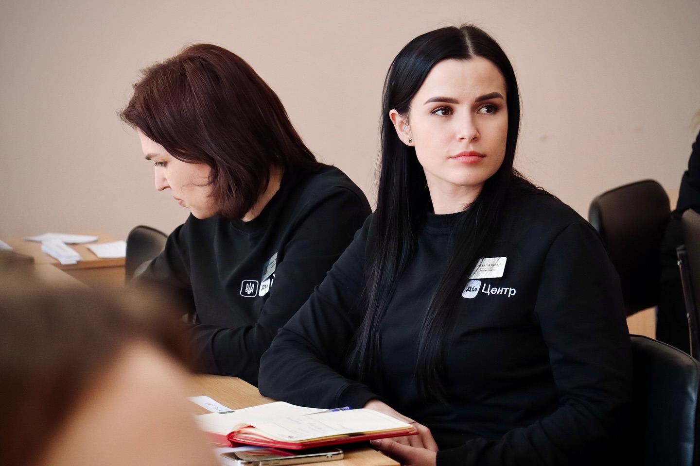 Дві жінки з темним волоссям та в чорних кофтах сидять за столом.