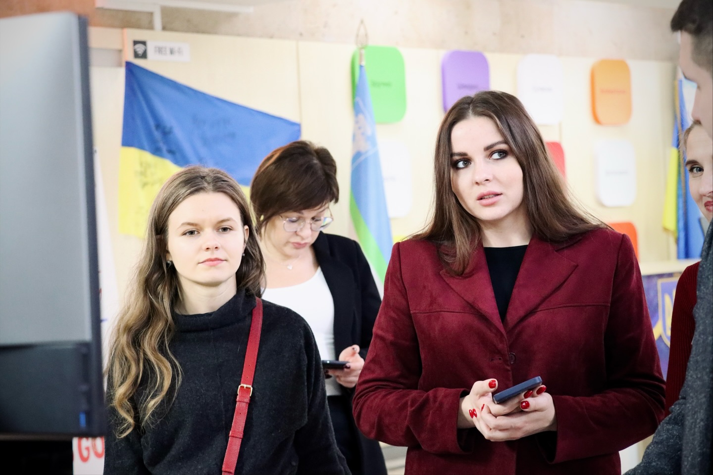 Кілька жінок у приміщенні, дві на передньому плані, на задньому плані — жінка з телефоном і прапори