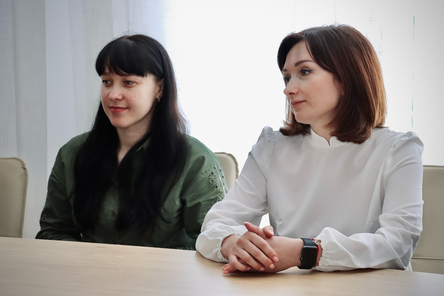 Дві жінки сидять за столом, на фоні — світлі штори.