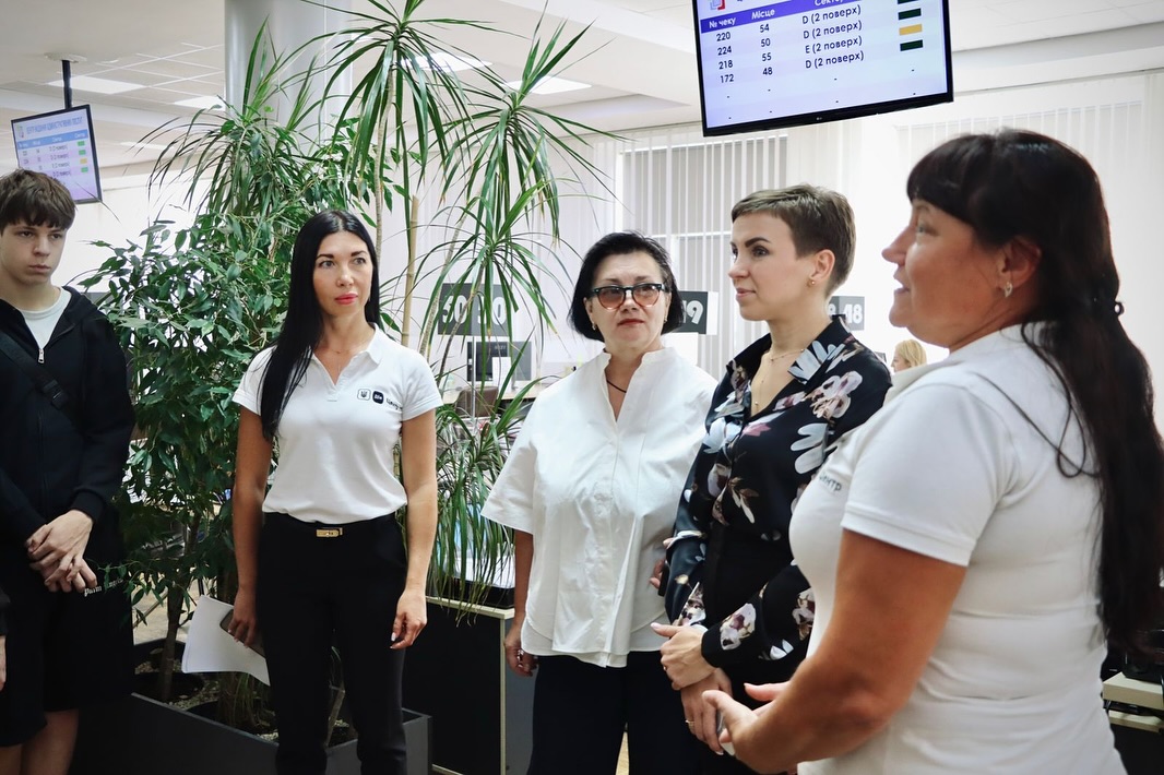 Група людей стоїть у приміщенні, поруч табло з номерами черги та зелені рослини.