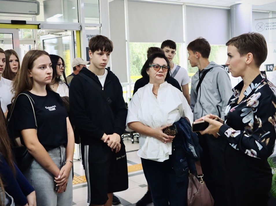 Група людей слухає жінку в темному одязі, яка тримає телефон і пояснює щось у приміщенні.