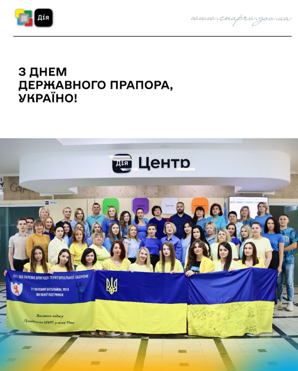 Колектив працівників у жовто-блакитному одязі тримає державний прапор України в приміщенні ЦНАП.