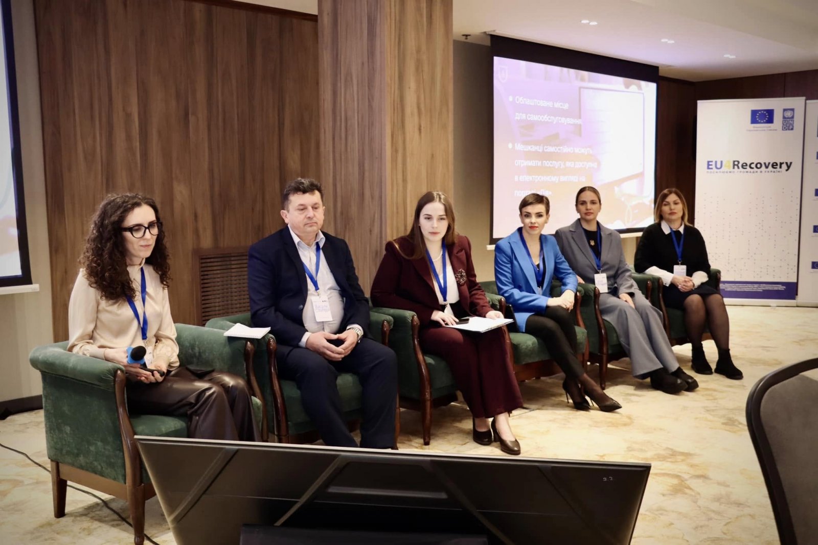 Шестеро спікерів у кріслах, одна з жінок тримає мікрофон. На задньому плані екран із презентацією.