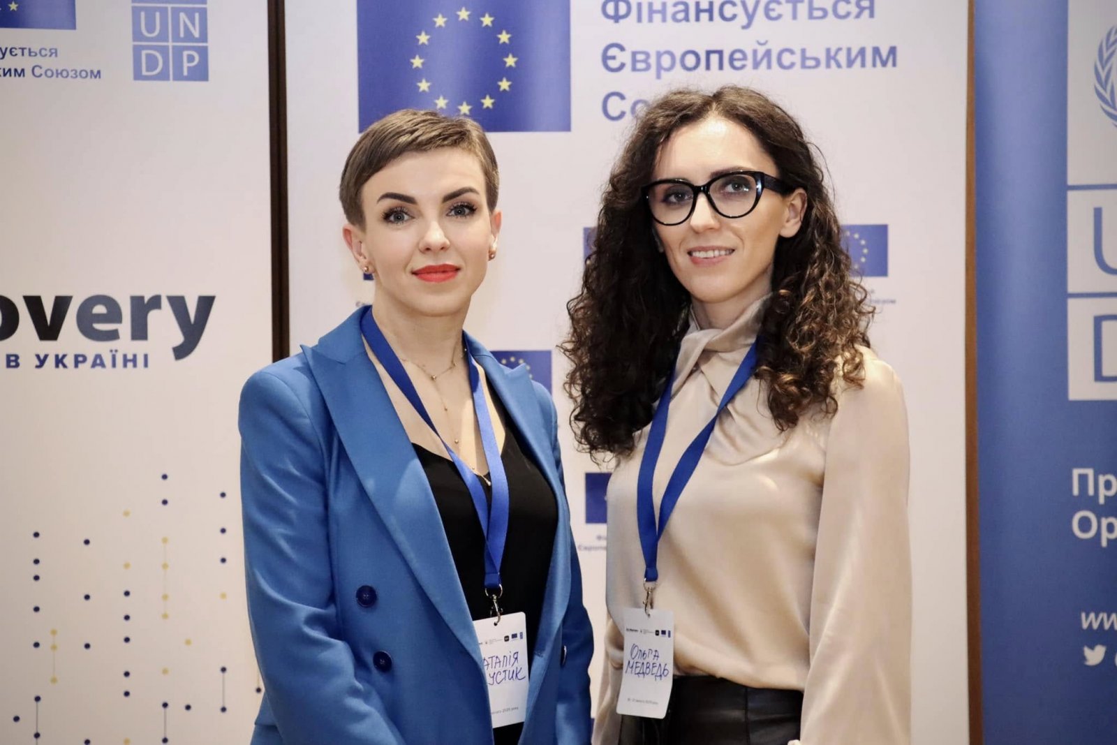 Дві жінки у діловому одязі з бейджами позують на тлі банерів.