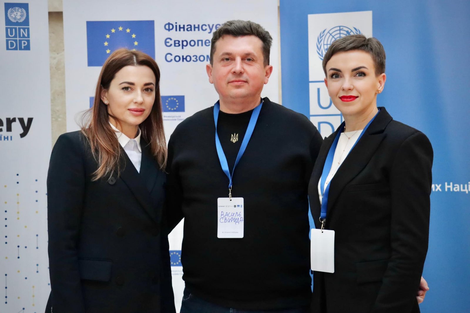 Дві жінки та чоловік з бейджами позують на тлі банерів.