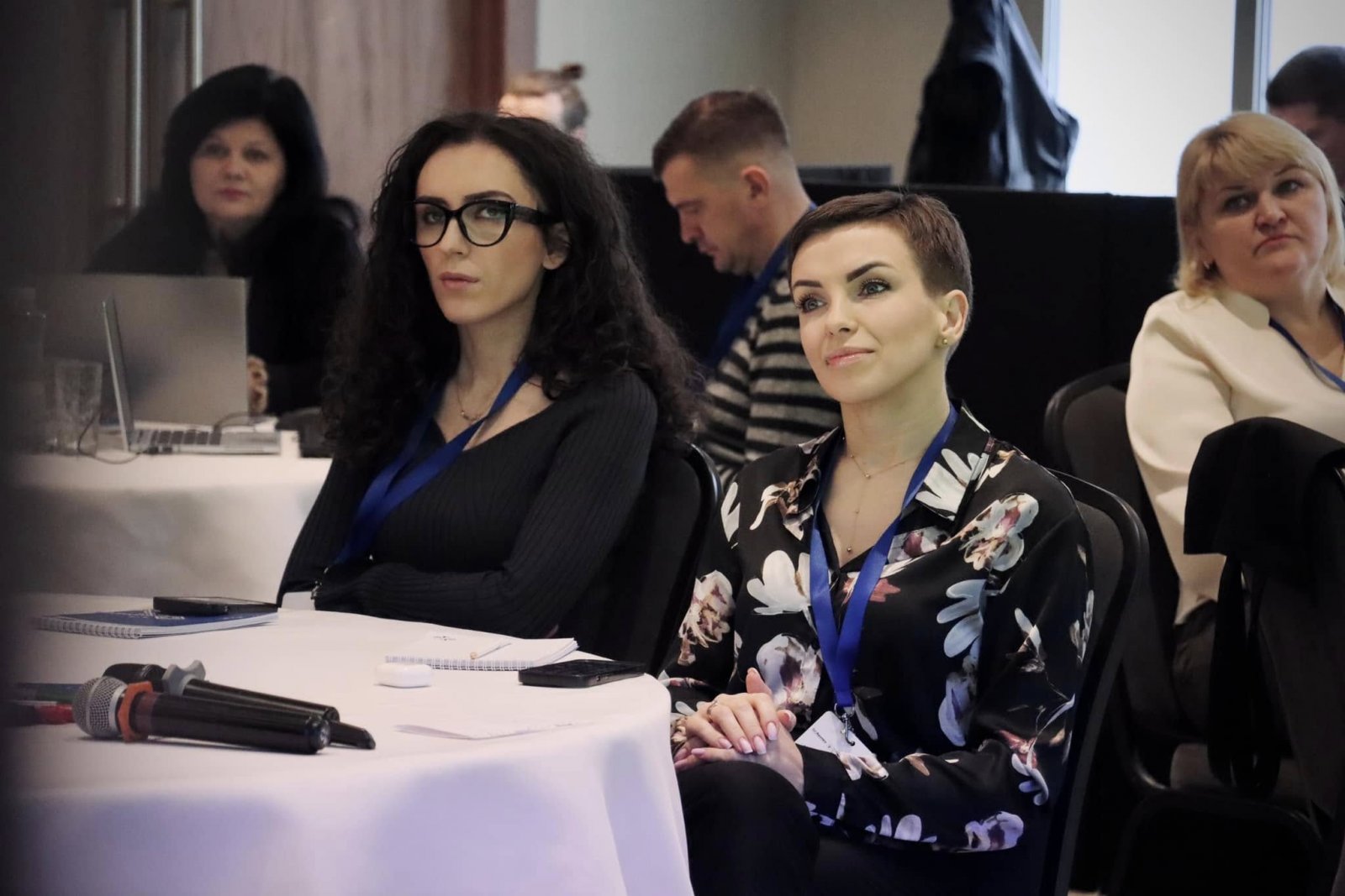 Дві жінки слухають виступ заході. Вони сидять за столом із блокнотами та мікрофонами.