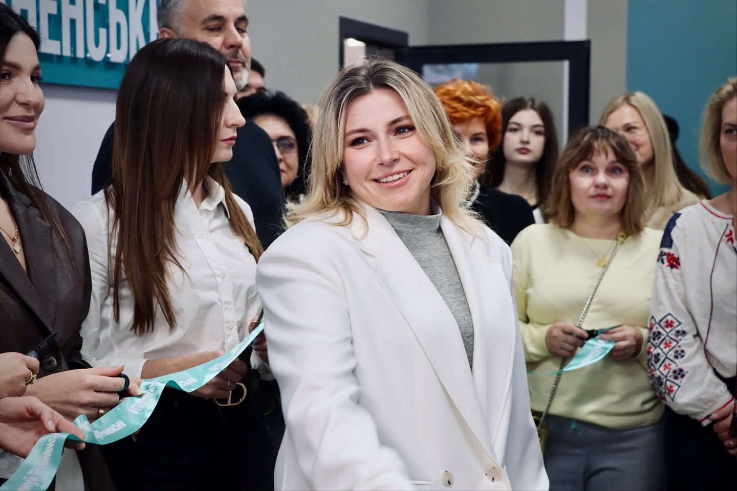 Жінка у білому одязі посміхається, на задньому фоні - багато людей.