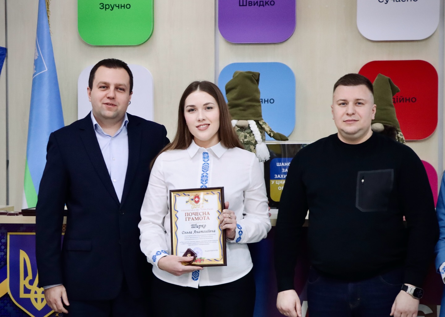 Дівчина тримає грамоту, біля неї стоять двоє чоловіків.