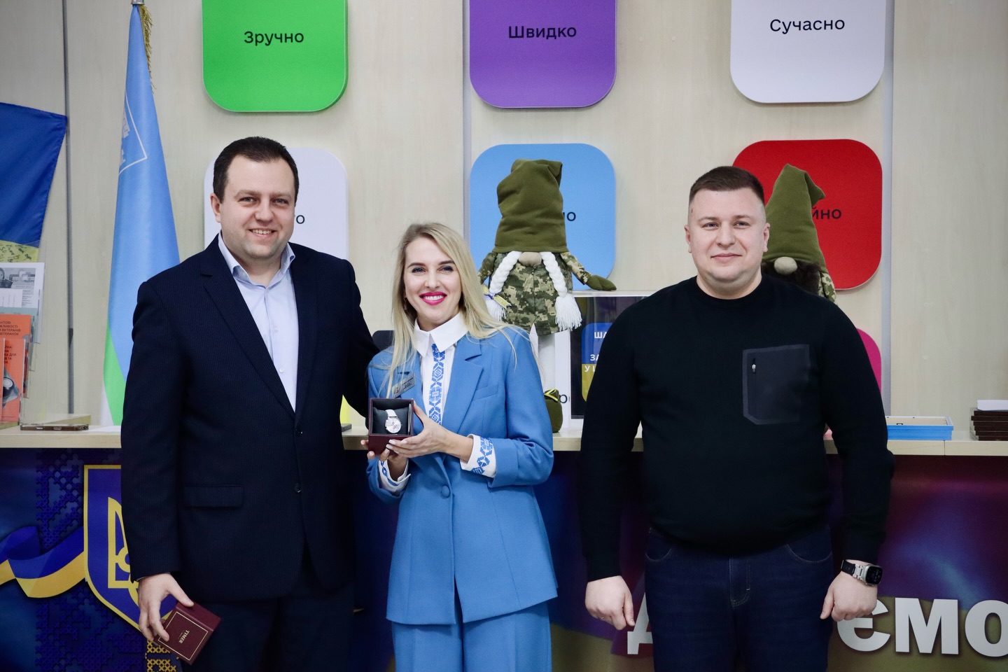 Жінка тримає годинник та стоїть між двома чоловіками.