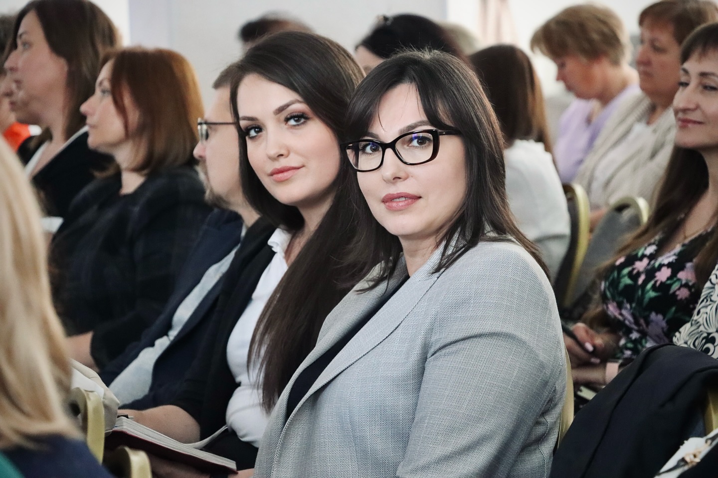 Група учасників семінару, зосереджено слухають виступ. Дві жінки на передньому плані дивляться в камеру