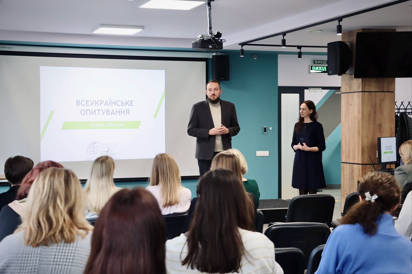 Чоловік стоїть перед людьми, які сидять у приміщенні.