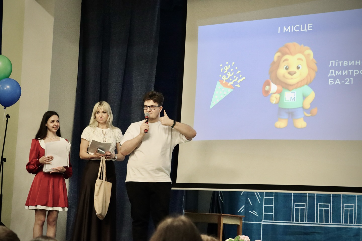 Хлопець з мікрофоном та дві жінки на сцені перед екраном.
