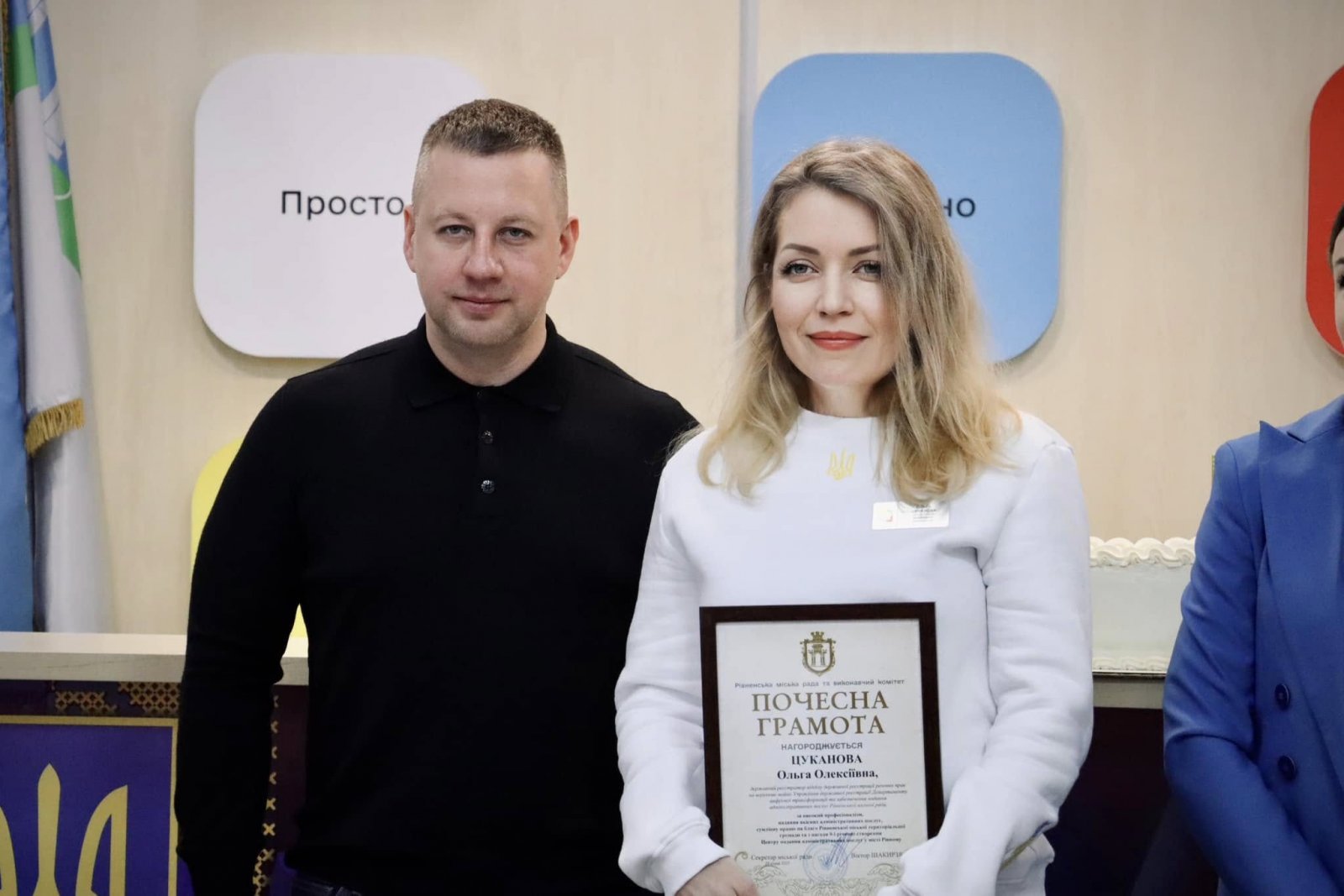 Чоловік та жінка у приміщені  ЦНАПу. Жінка тримає у руках почесну грамоту.