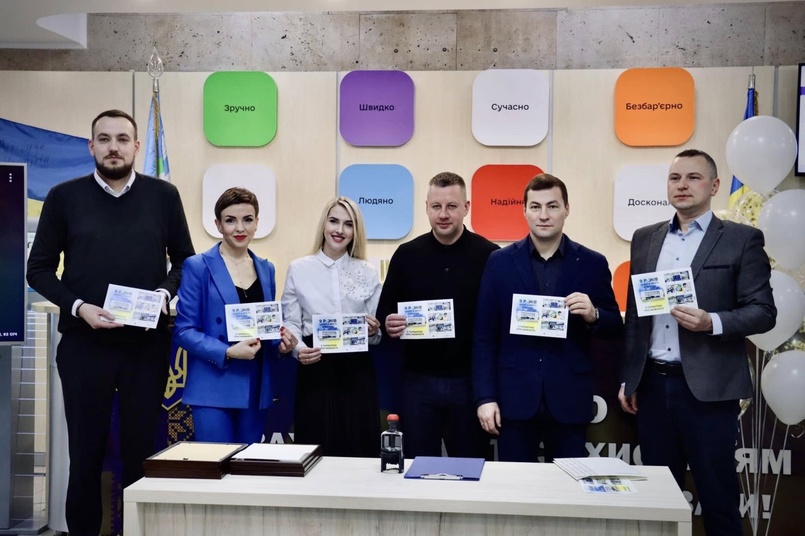 Чоловіки та жінки стоять у приміщені  ЦНАПу в місті Рівному та тримають у руках марки.
