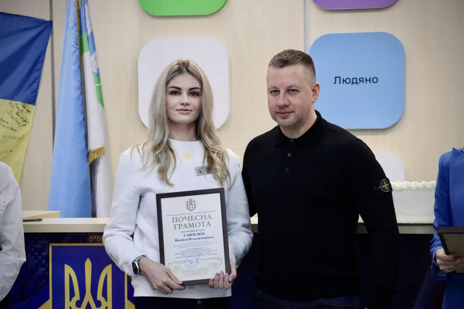 Чоловік та жінка стоять у приміщені  ЦНАПу в місті Рівному. Жінка тримає у руках почесну грамоту.