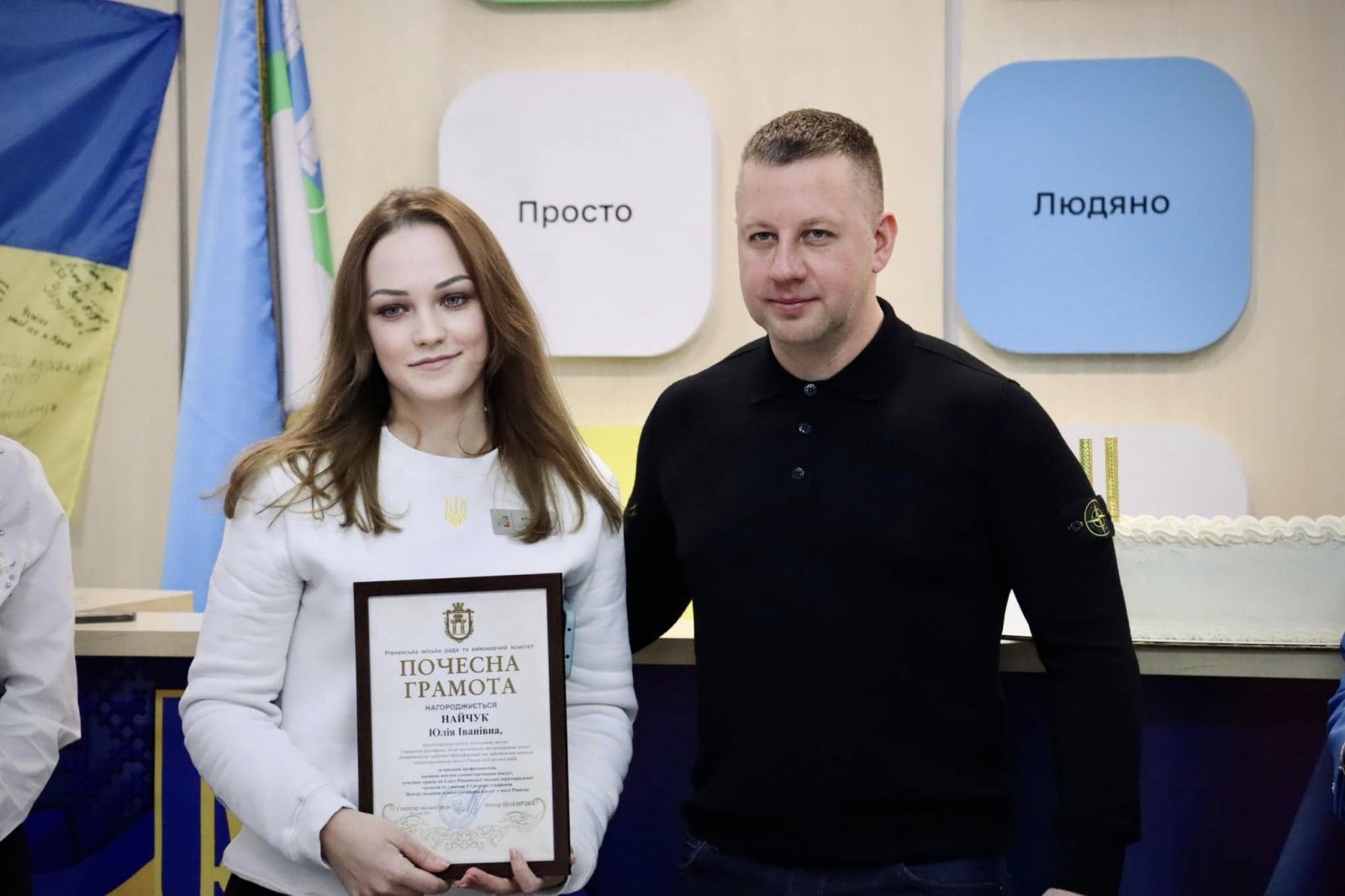 Чоловік та жінка стоять у приміщені  ЦНАПу в місті Рівному. Жінка тримає почесну грамоту.