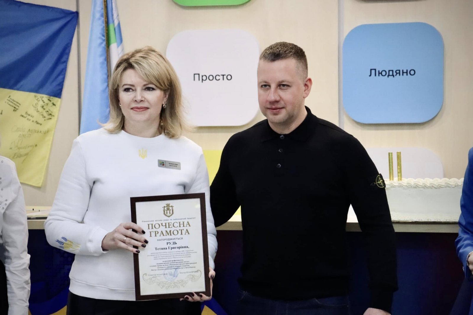 Чоловік та жінка перебувають у приміщені  ЦНАПу. Жінка тримає у руках почесну грамоту.