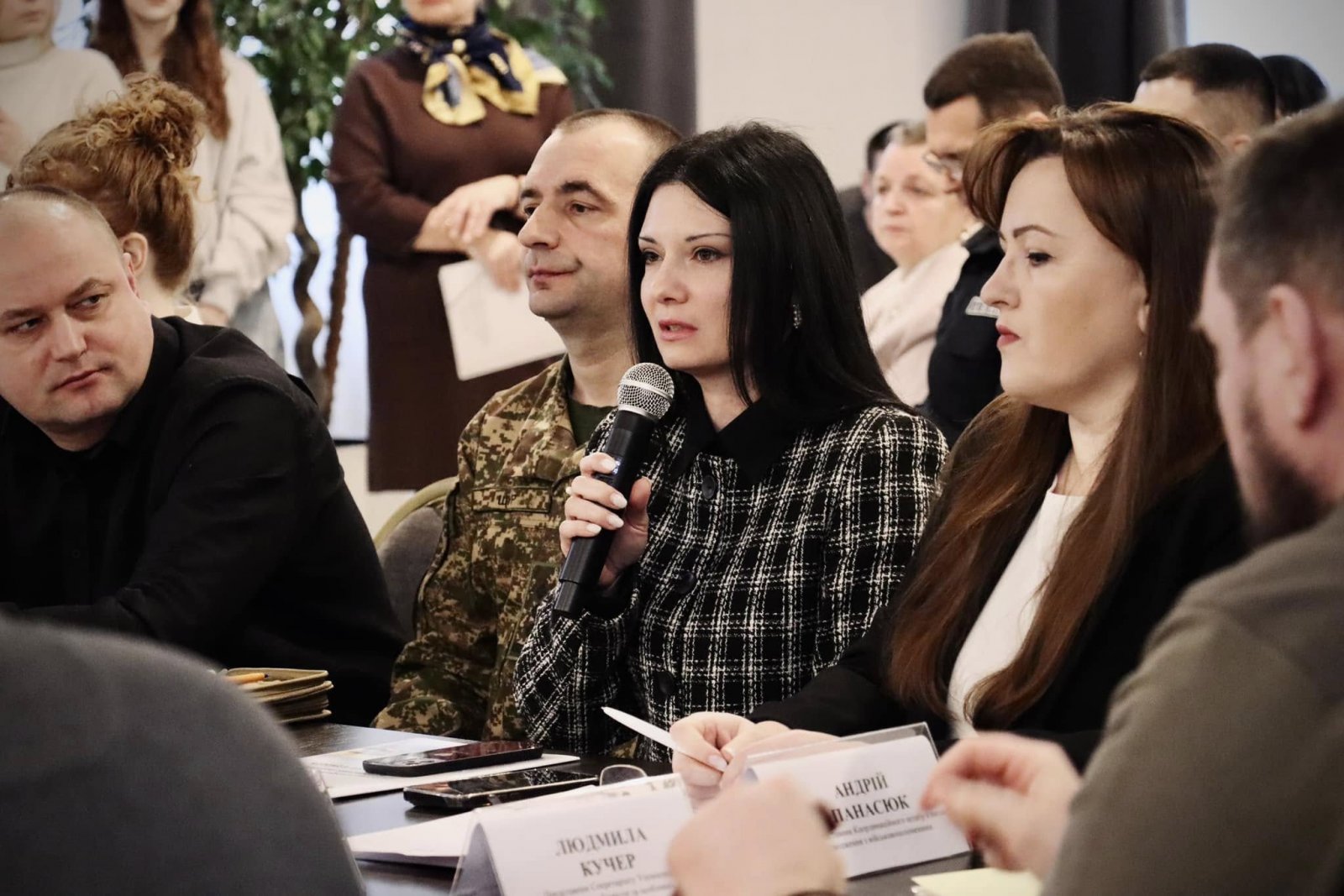 Жінка сидить за столом та тримає у руках мікрофон. На задньому фоні інші учасники зустрічі.