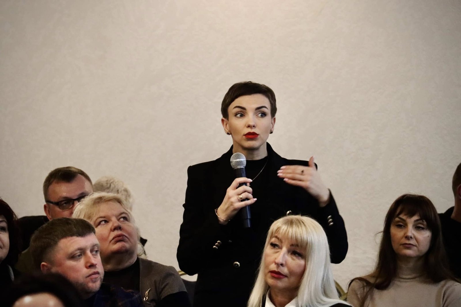 Жінка тримає у руках мікрофон та проголошує доповідь. Інші присутні її слухають.