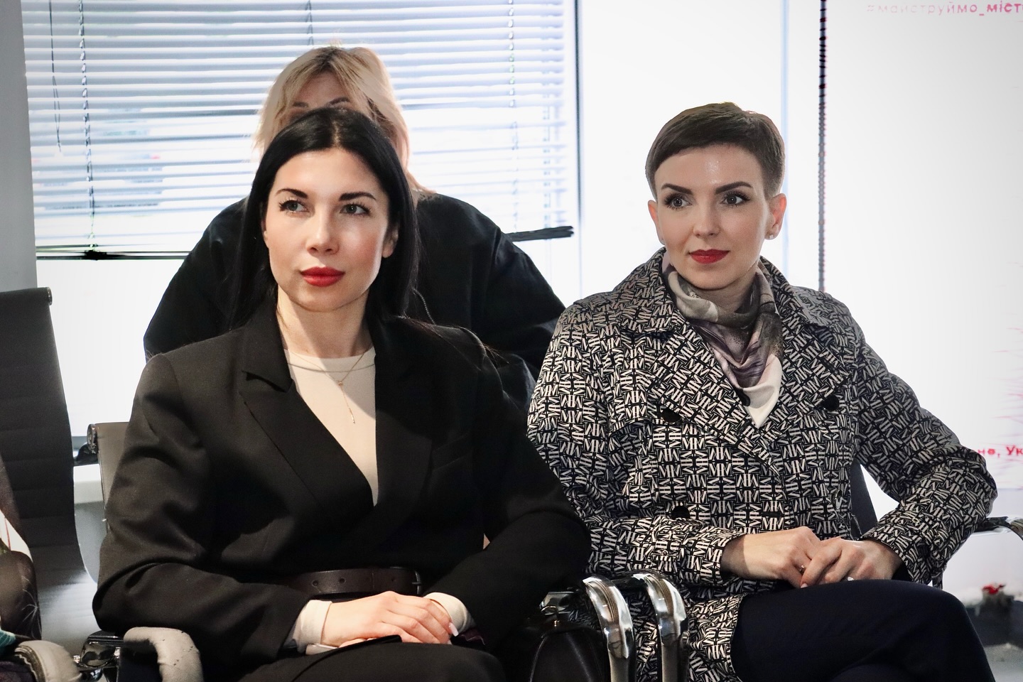 Дві жінки у верхньому одязі сидять у приміщенні.