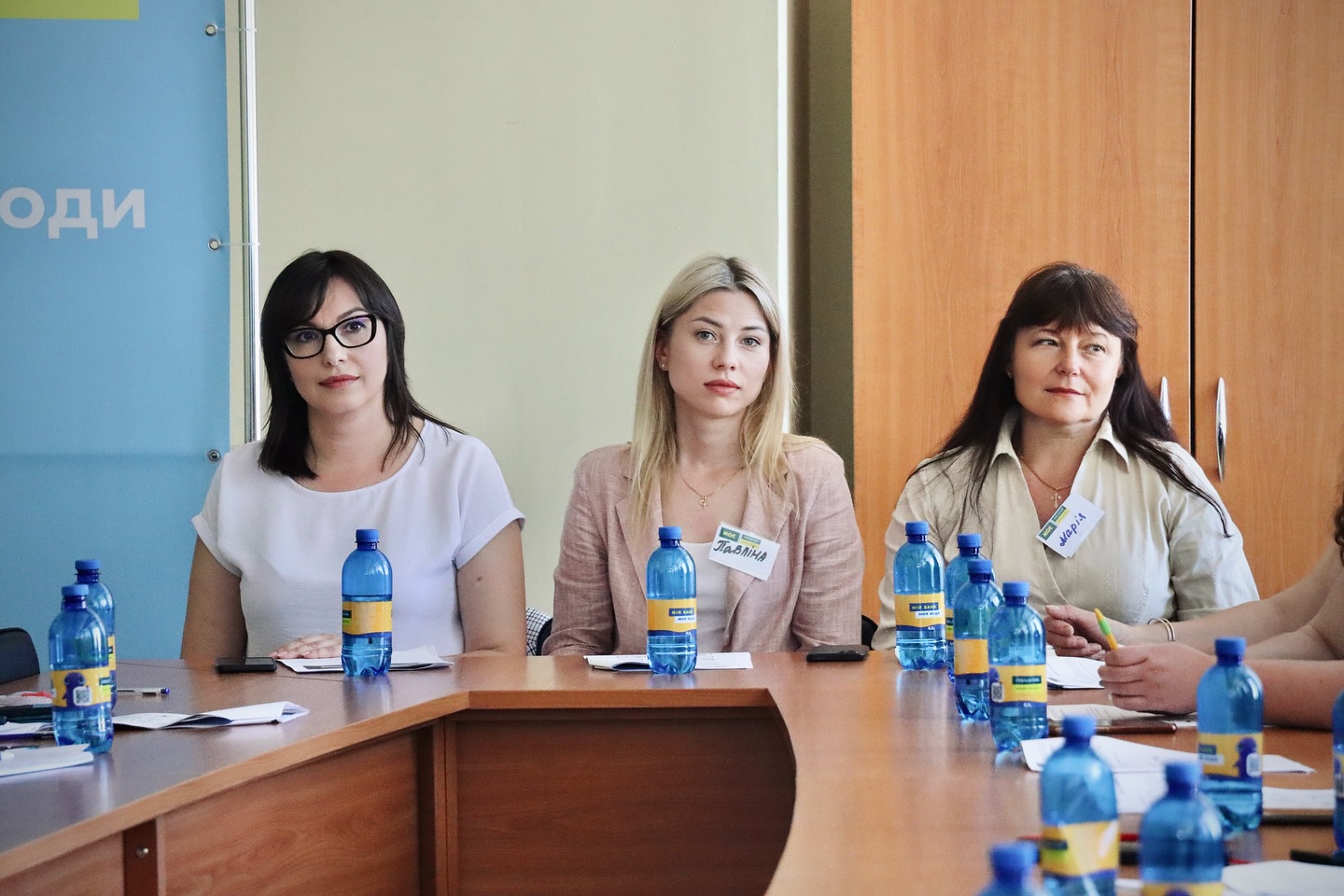 Три жінки сидять за круглим конференц-столом у залі засідань, перед ними – пляшки з водою, блокноти та бейджі.